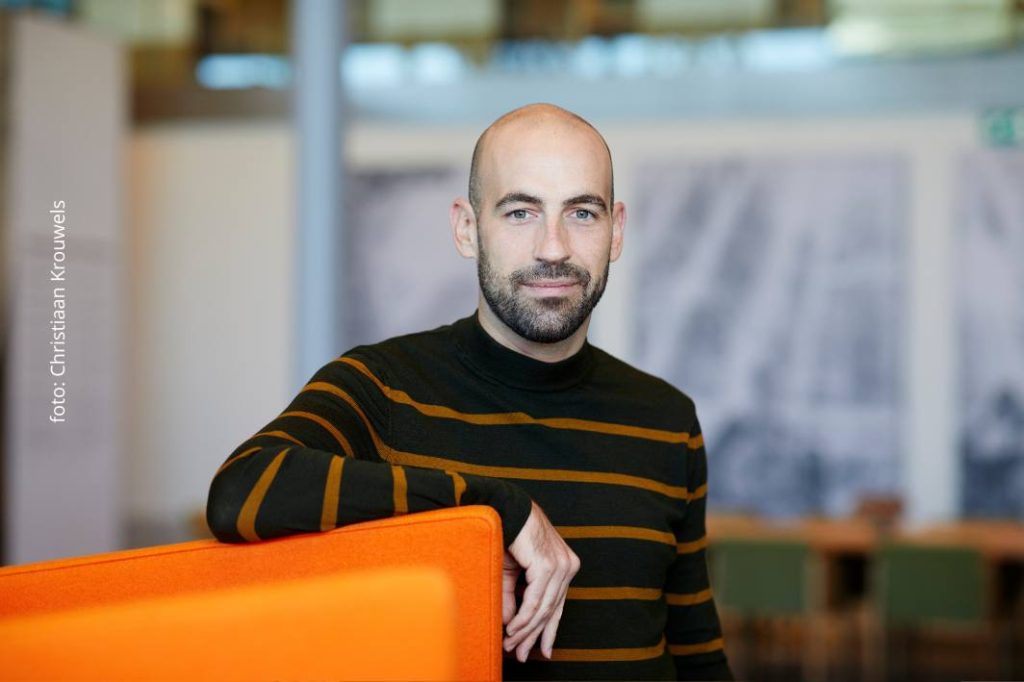 Man met zwarte trui met oranje strepen leunt met zijn arm op een oranje stoel en kijkt recht in de camera