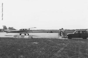 Een zwart-wit foto van een vliegtuig op de airstrip in Drachten dat in opdracht van Philips werd aangelegd. Aan de rand van de airstrip zijn mensen uit een oude Renault gestapt. 