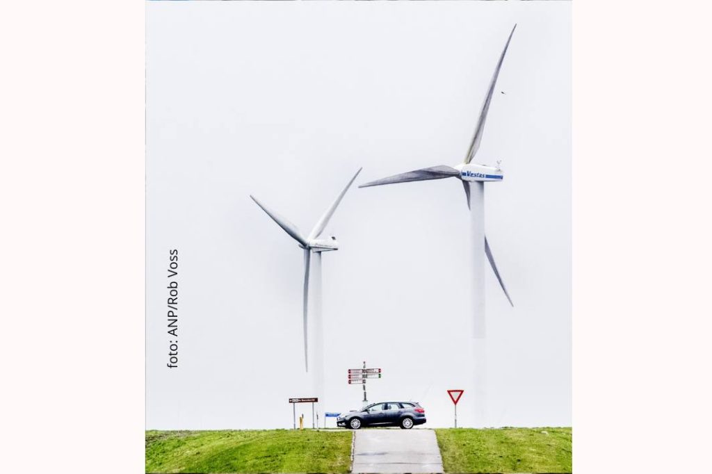 Een personenauto staat op een dijk met op de achtergrond twee heel grote windmolens