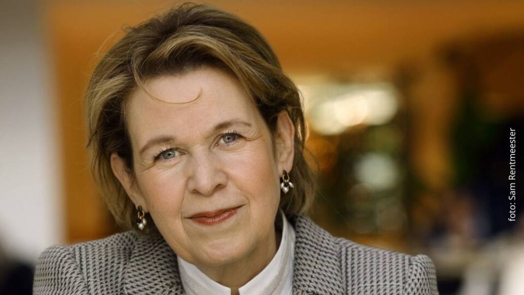 Portret van Edith Nordmann, India-expert, die recht in de camera kijkt en licht glimlacht, gekleed in een lichtgekleurde blouse en een geruit jasje, met een warme, onscherpe achtergrond binnenshuis.