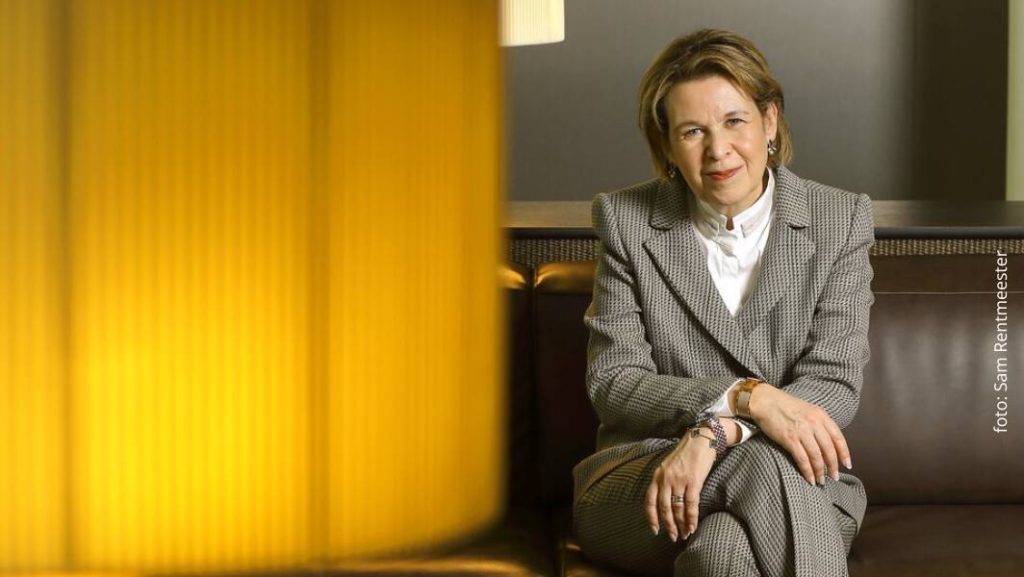 Edith Nordmann, India-expert, zit op een bank en kijkt recht in de camera, gekleed in een geruit pak, met een warme, sfeervolle binnenruimte.